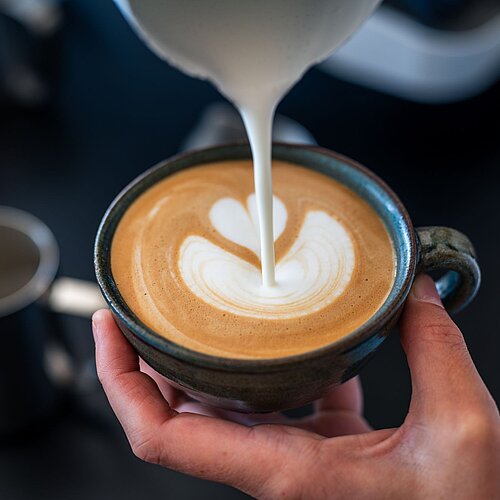 Hand gießt Milch in eine Cappuccino-Tasse und kreiert eine Blume aus Milchschaum