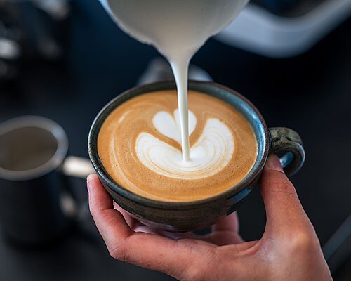 Hand gießt Milch in eine Cappuccino-Tasse und kreiert eine Blume aus Milchschaum