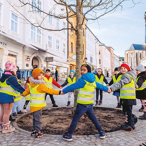 Kinder stehen um einen neu gepflanzten Baum und halten sich an ihren Händen