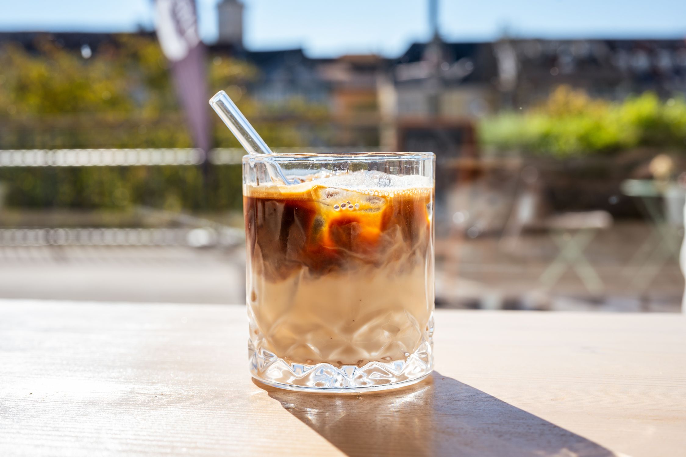 Glas mit Café Latte und Glasstrohhalm, im Hintergrund Blick auf die Villacher Innenstadt